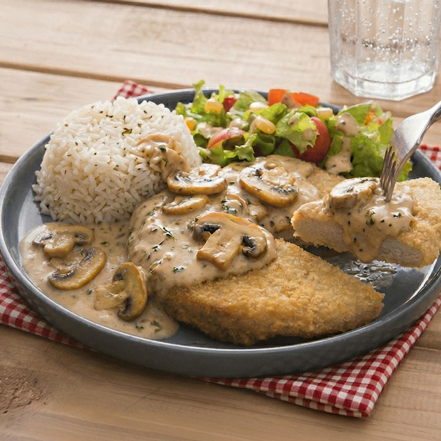 Plato de arroz con milanesa de pollo apanada bañada en salsa cremosa de champiñones, acompañado de ensalada fresca y vaso con bebida fría sobre mesa de madera